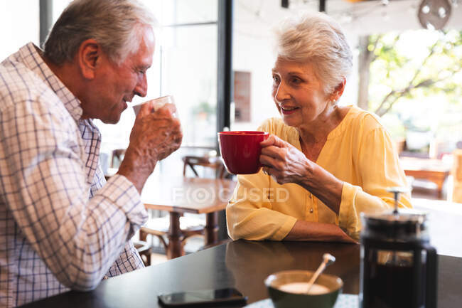 Una pareja caucásica jubilada en casa sentada en la encimera de la isla en su cocina, hablando, sonriendo y tomando café juntos en un día soleado, pareja aislada durante la pandemia de coronavirus covid19 - foto de stock