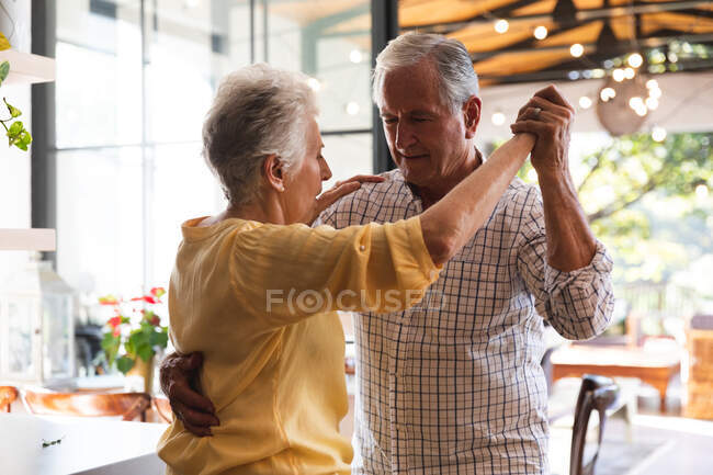 Glückliche Rentner kaukasischen Paar zu Hause Händchen haltend, tanzen zusammen in ihrer Küche und lächeln, zu Hause zusammen isolieren während Coronavirus covid19 Pandemie — Stockfoto