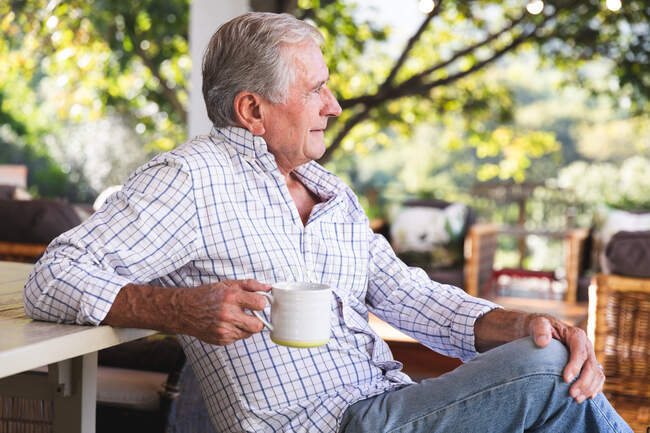 Вид сбоку счастливого пожилого кавказца на пенсии дома в саду возле его дома в солнечный день, сидящего на скамейке, держащего чашку кофе, глядящего в сторону и улыбающегося, самоизолирующегося во время пандемии коронавируса — стоковое фото