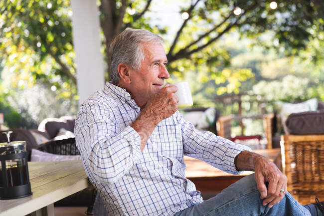 Вид сбоку счастливого пожилого кавказца на пенсии дома в саду возле его дома в солнечный день, сидящего на скамейке и пьющего чашку кофе, отворачивающегося и улыбающегося, самоизолирующегося во время пандемии коронавируса — стоковое фото