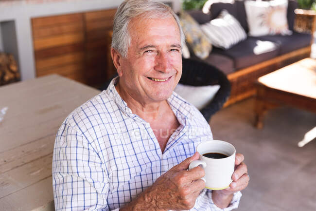 Portrait d'un homme caucasien âgé à la retraite heureux à la maison dans son salon par une journée ensoleillée, assis et buvant une tasse de café, regardant vers la caméra et souriant, auto-isolant pendant la covid19 coronavirus pandémie — Photo de stock