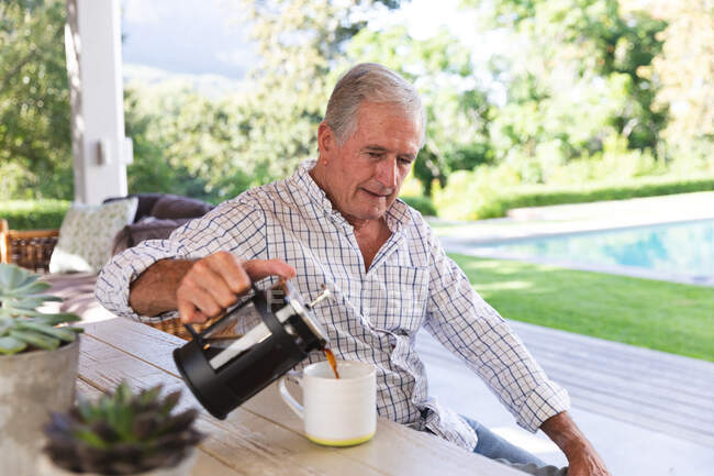 Seitenansicht eines glücklichen Rentner kaukasischen Mann zu Hause im Garten vor seinem Haus an einem sonnigen Tag, sitzt auf einer Bank an einem Tisch und gießt eine Tasse Kaffee und lächelt, selbstisolierend während Coronavirus covid19 Pandemie — Stockfoto