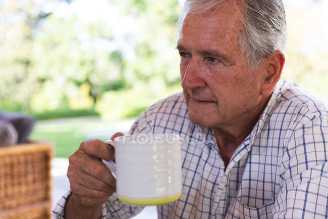 Gros plan d'un homme caucasien âgé à la retraite heureux à la maison dans le jardin à l'extérieur de sa maison par une journée ensoleillée, assis sur un banc, tenant une tasse de café, regardant loin et souriant, auto-isolant pendant la pandémie de coronavirus covid19 — Photo de stock
