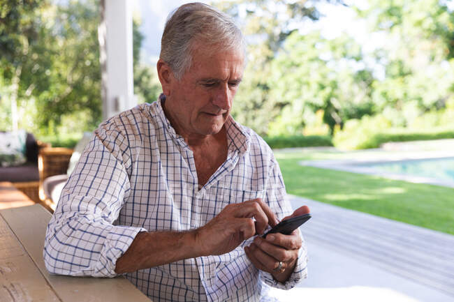 Hombre caucásico mayor guapo disfrutando de su retiro, sentado en una mesa en el jardín en el sol mensajería de texto con un teléfono móvil, auto aislamiento durante coronavirus covid19 pandemia - foto de stock