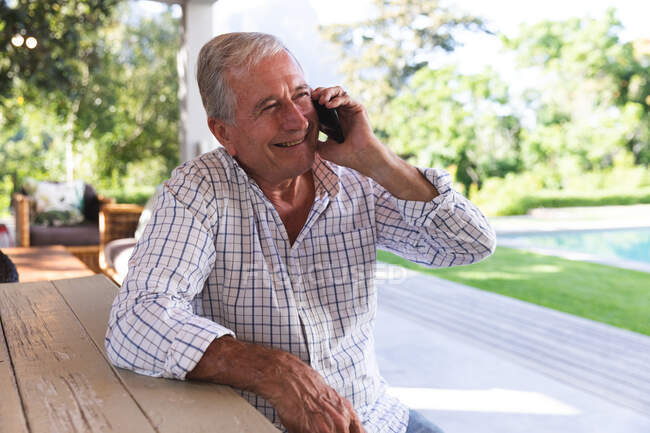 Charmant homme caucasien âgé profitant de sa retraite, assis à une table dans le jardin au soleil parlant sur un téléphone portable, auto-isolant pendant la pandémie de coronavirus covid19 — Photo de stock