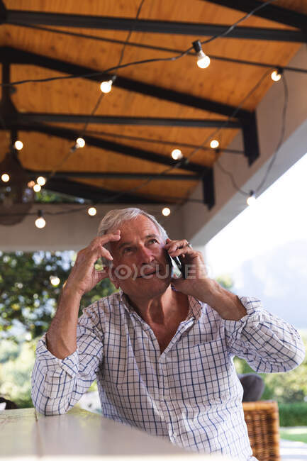 Bell'uomo caucasico anziano che si gode la pensione, seduto a un tavolo in giardino sotto una terrazza sul tetto al sole a parlare su un telefono cellulare, autoisolante durante la pandemia coronavirus covid19 — Foto stock