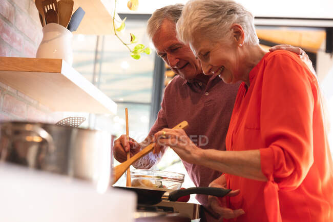 Felice pensionato coppia caucasica anziani a casa, preparando il cibo nella loro cucina, sia padelle mescolando sul piano cottura e sorridente, a casa insieme isolando durante il coronavirus covid19 pandemia — Foto stock