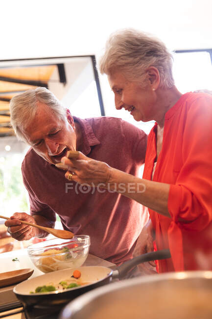 Счастливая пожилая кавказская пара дома, готовящая еду на своей кухне вместе, женщина, дающая мужчине полный рот еды из деревянной ложки, дома вместе изолирующая во время пандемии коронавируса — стоковое фото