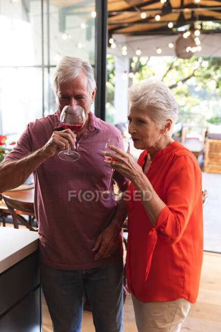 Feliz pareja de ancianos caucásicos jubilados en casa en su cocina, de pie y bebiendo vasos de vino y ambos sonriendo, en casa juntos aislando durante coronavirus covid19 pandemia - foto de stock