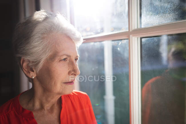 Gros plan d'une femme caucasienne âgée à la retraite à la maison dans son salon, regardant par la fenêtre par une journée ensoleillée et pensant, s'isolant pendant une pandémie de coronavirus covid19 — Photo de stock