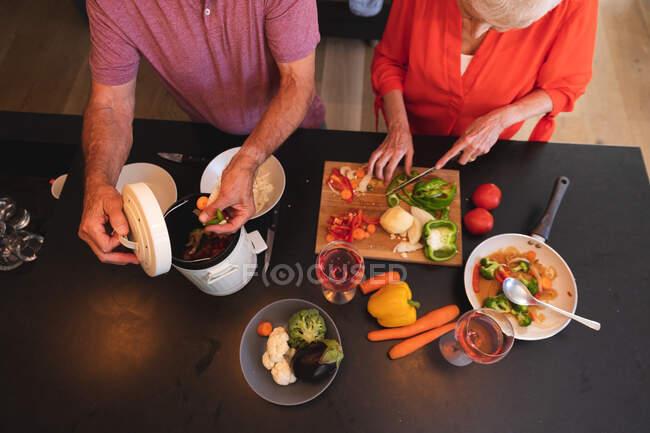 Vue aérienne d'un couple caucasien âgé à la retraite à la maison, préparant la nourriture dans leur cuisine, la femme coupant des légumes sur une planche à découper et l'homme mettant les déchets dans un récipient de compostage — Photo de stock