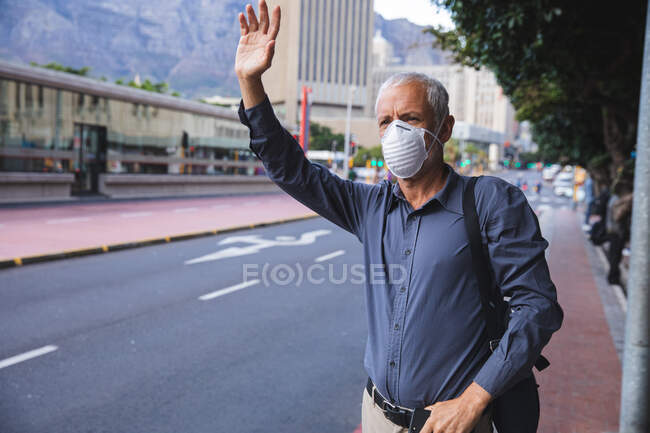 Homem caucasiano sênior nas ruas da cidade durante o dia, usando uma máscara facial contra o coronavírus, vívido 19, saudando um táxi . — Fotografia de Stock