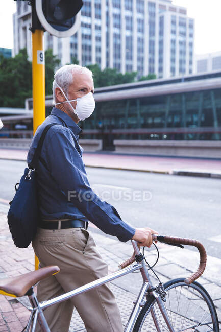 Senior Caucasian man out and about in the city streets during the day, wearing a face mask against coronavirus, covid 19, wheeling his bicycle. — Stock Photo