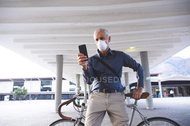 Hombre caucásico mayor por las calles de la ciudad durante el día, con una máscara facial contra el coronavirus, covid 19, sentado en su bicicleta y usando un teléfono inteligente. - foto de stock