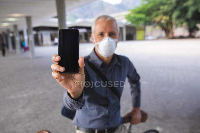 Uomo anziano caucasico in giro per le strade della città durante il giorno, indossando una maschera contro il coronavirus, covid 19, seduto sulla sua bicicletta e mostrando uno smartphone alla fotocamera. — Foto stock