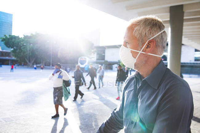 Hombre caucásico mayor fuera y alrededor de las calles de la ciudad durante el día, con una máscara facial contra el coronavirus, covid 19 con otras personas caminando en el fondo. - foto de stock