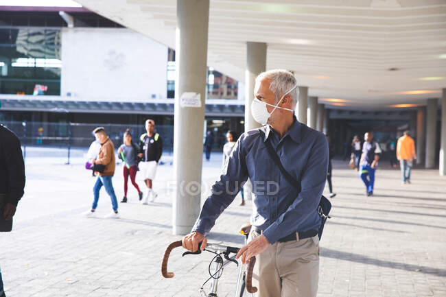 Senior Caucasian man out and about in the city streets during the day, wearing a face mask against coronavirus, covid 19, wheeling a bicycle. — Stock Photo