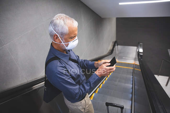Homme caucasien âgé, portant un masque facial contre le coronavirus, covide 19, debout sur un escalier roulant dans une station de métro et utilisant son smartphone. — Photo de stock