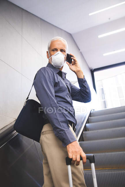 Senior Caucasian man , wearing a face mask against coronavirus, covid 19, using an escalator in a metro station, talking on his smartphone, and pulling a suitcase. — Stock Photo