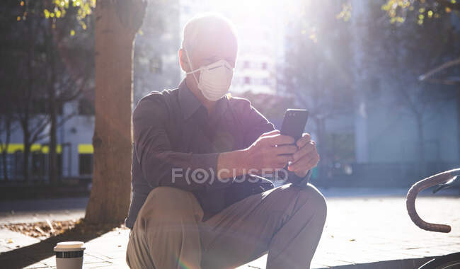 Hombre caucásico mayor fuera y alrededor de las calles de la ciudad durante el día, con una máscara facial contra el coronavirus, covid 19, sentado en las escaleras y utilizando su teléfono inteligente. - foto de stock