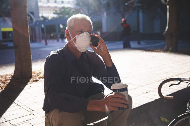 Homem caucasiano sênior para fora e aproximadamente nas ruas da cidade durante o dia, usando uma máscara facial contra coronavírus, covid 19, sentado nas escadas, segurando uma xícara de café takeaway e usando seu smartphone . — Fotografia de Stock