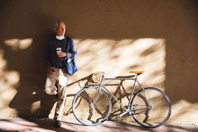 Homme caucasien âgé dehors et autour dans les rues de la ville pendant la journée, portant un masque facial contre le coronavirus, covid 19, appuyé sur le mur et tenant une tasse de café à emporter pendant que son vélo est debout à côté de lui. — Photo de stock