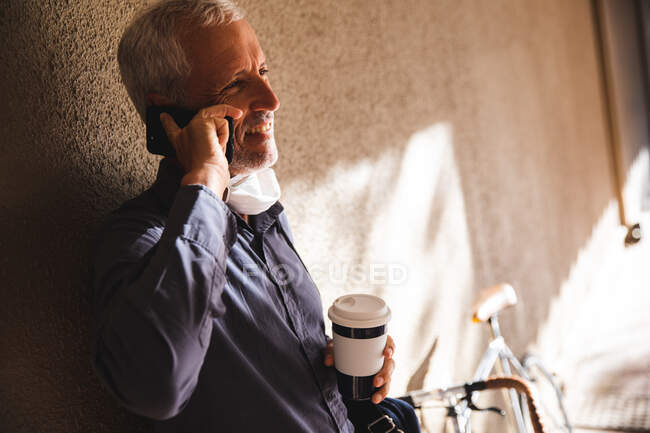Homme caucasien âgé dehors et autour dans les rues de la ville pendant la journée, portant un masque facial contre le coronavirus, covid 19, appuyé sur le mur et boire du café à emporter pendant que son vélo est debout à côté de lui. — Photo de stock
