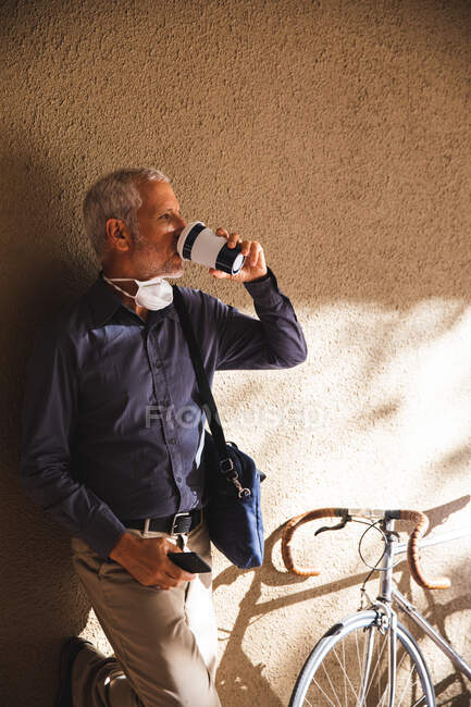 Senior Caucasian man out and about in the city streets during the day, wearing a face mask against coronavirus, covid 19, leaning on the wall and drinking takeaway coffee while his bicycle is standing next to him. — Stock Photo