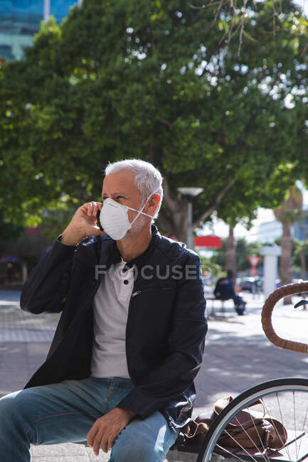 Hombre caucásico mayor por las calles de la ciudad durante el día, con una máscara facial contra el coronavirus, covid 19, sentado en un banco y usando su teléfono inteligente. - foto de stock