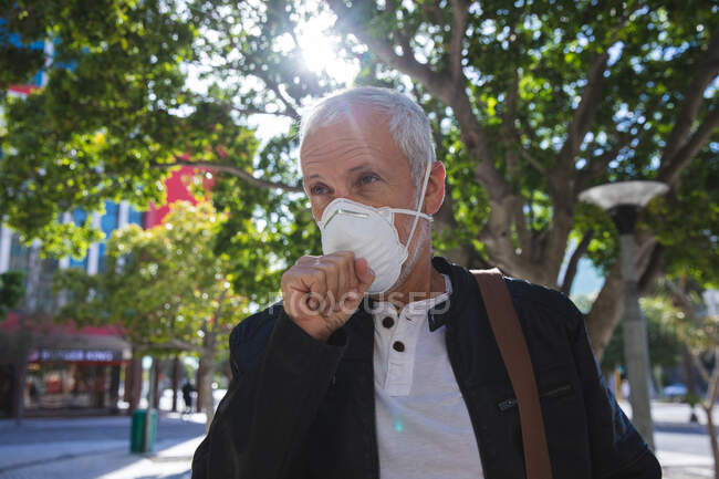 Uomo anziano caucasico in giro per le strade della città durante il giorno, indossando una maschera contro il coronavirus, covid 19, coprendo il suo viso mentre tossisce. — Foto stock