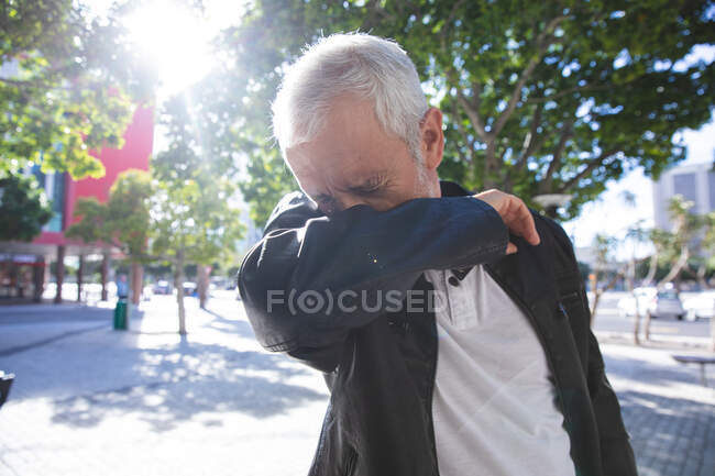 Hombre caucásico mayor con ropa casual, fuera y alrededor en las calles de la ciudad durante el día, cubriéndose la cara mientras tose. - foto de stock