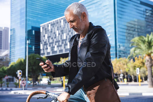 Homme caucasien âgé dans les rues de la ville pendant la journée, assis sur son vélo et en utilisant un smartphone. — Photo de stock