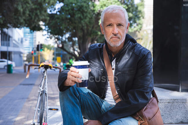 Älterer Mann aus dem Kaukasus, lässig gekleidet, tagsüber auf den Straßen der Stadt unterwegs, sitzt auf der Treppe, hält eine Tasse Kaffee zum Mitnehmen in der Hand und blickt direkt in die Kamera. — Stockfoto
