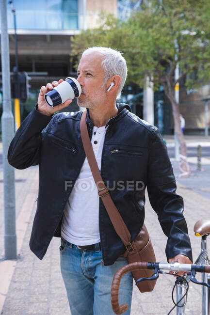 Senior Caucasian man, wearing earphones, out and about in the city streets during the day, drinking takeaway coffee and wheeling a bicycle. — Stock Photo