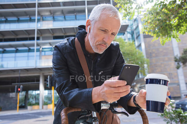 Homme caucasien âgé, portant des écouteurs, dans les rues de la ville pendant la journée, tenant un café à emporter et utilisant son smartphone tout en s'appuyant sur un vélo. — Photo de stock