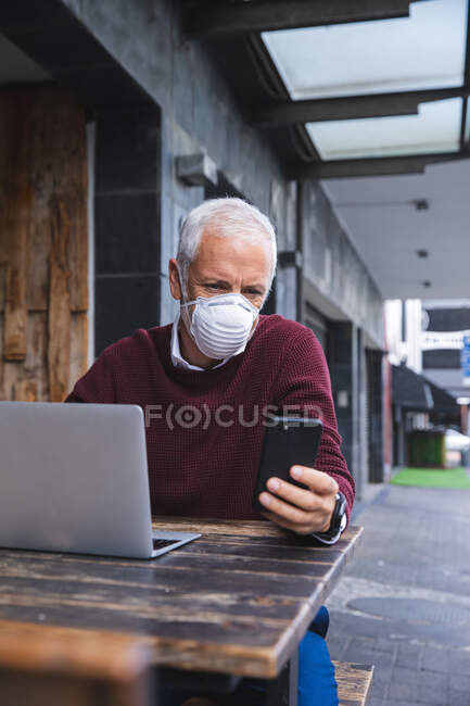 Старший кавказський чоловік сидить за столом на терасі кави, одягнувши маску обличчя проти коронавірусу, ковидка 19, за допомогою смартфона і ноутбука.. — стокове фото