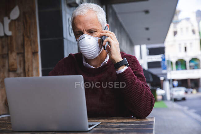 Uomo anziano caucasico seduto a un tavolo in una terrazza di caffè, indossando una maschera contro il coronavirus, covid 19, utilizzando uno smartphone e un computer portatile. — Foto stock