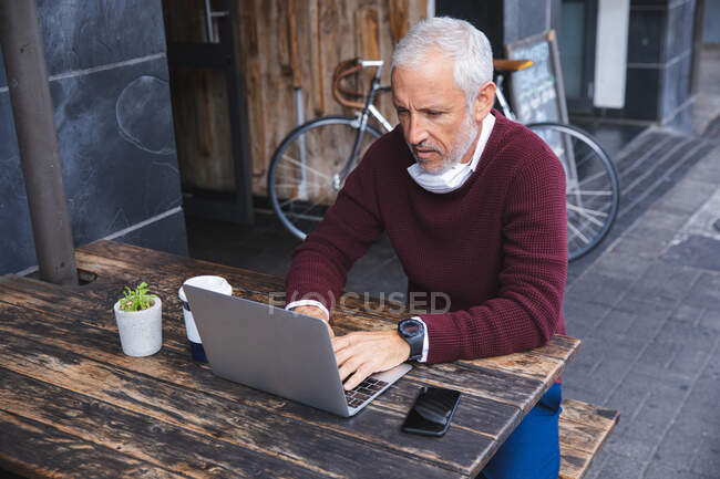 Hombre caucásico mayor sentado en una mesa en una terraza de café, con una máscara facial contra coronavirus, covid 19, usando un teléfono inteligente y un ordenador portátil. - foto de stock