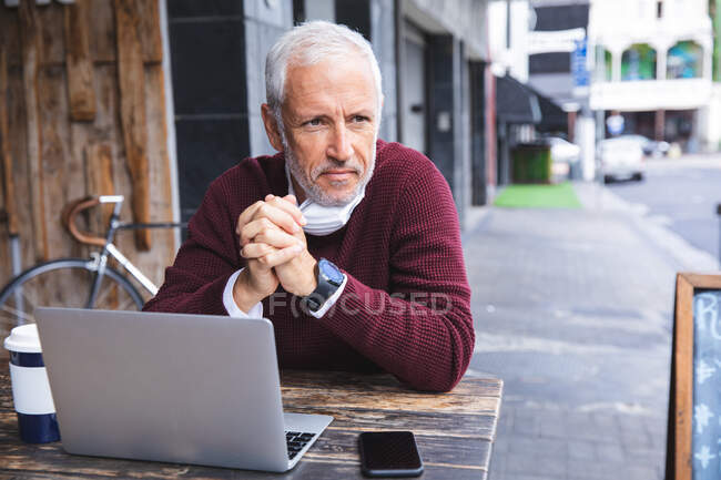 Homem caucasiano sênior sentado em uma mesa em um terraço de café, usando uma máscara facial contra coronavírus, covid 19, usando um smartphone e computador portátil . — Fotografia de Stock