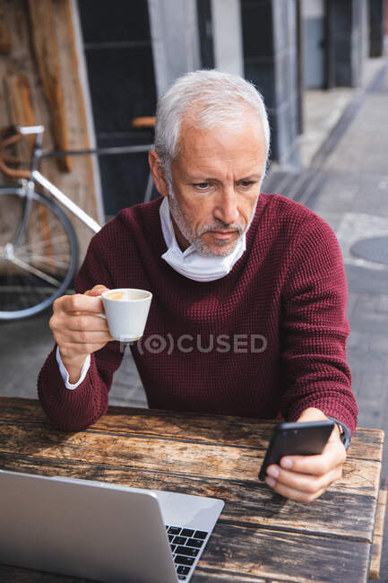 Uomo anziano caucasico seduto a un tavolo in una terrazza di caffè, indossando una maschera contro il coronavirus, covid 19, bere caffè e utilizzando uno smartphone. — Foto stock