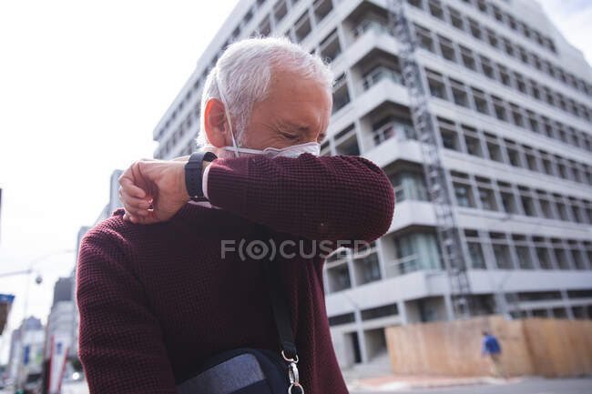 Uomo anziano caucasico in giro per le strade della città durante il giorno, indossando una maschera contro il coronavirus, covid 19, coprendo il suo viso mentre tossisce. — Foto stock