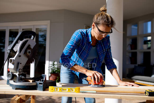 Caucasian woman spending time at home self isolating and social distancing in quarantine lockdown during coronavirus covid 19 epidemic, doing DIY in her garden, sanding wooden boards. — Stock Photo