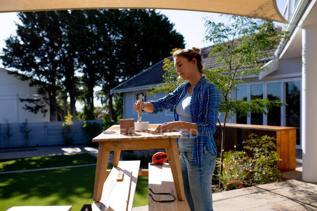 Caucasian woman spending time at home self isolating and social distancing in quarantine lockdown during coronavirus covid 19 epidemic, doing DIY in her garden, painting wooden table. — Stock Photo