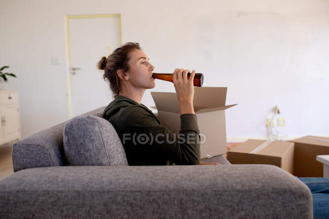 Кавказька жінка проводить вдома самоізоляцію і соціальну дистанцію в карантині під час епідемії коронавірусу covid 19, роблячи перерву, відпочиваючи на дивані і випивши пиво.. — стокове фото