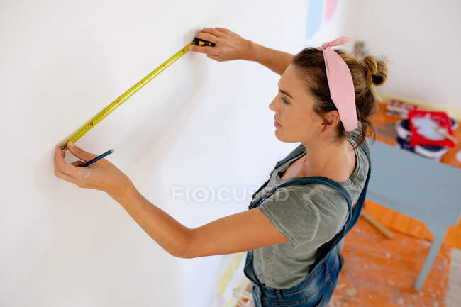 Caucasian woman wearing blue jeans dungarees, spending time at home self isolating and social distancing in quarantine lockdown during coronavirus covid 19 epidemic, measuring the walls. — Stock Photo