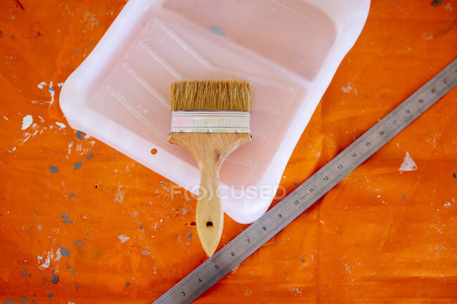 Top view of a paint brush, paint bucket and a ruler, lay on a table covered with orange foil, during DIY while social distancing and self isolation in quarantine lockdown during coronavirus covid 19 epidemic. — Stock Photo