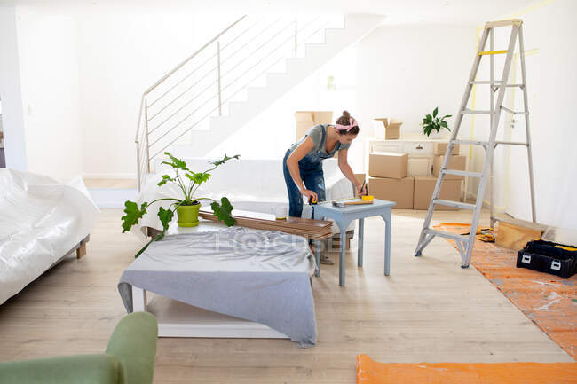 Caucasian woman wearing blue jeans dungarees, spending time at home self isolating and social distancing in quarantine lockdown during coronavirus covid 19 epidemic, tinkering a table. — Stock Photo