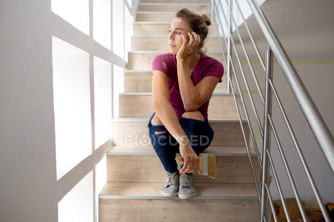 Кавказская женщина проводит время дома, изолируя себя и отдаляясь от общества в карантинной изоляции во время эпидемии коронавируса ковида 19, делая перерыв во время ремонта своего дома, сидя на лестнице и держа кисть. — стоковое фото