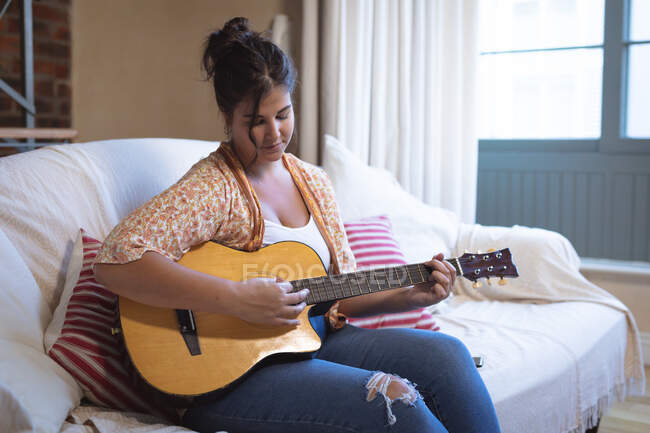 Vlogger mujer caucásica en casa en su sala de estar, sentado en un sofá y tocando la guitarra. Distanciamiento social y autoaislamiento en cuarentena. - foto de stock