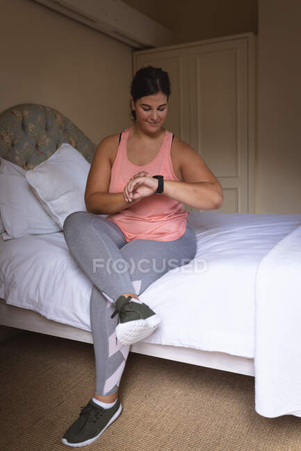 Vlogger mujer caucásica en casa en su dormitorio, preparándose para demostrar ejercicios para su blog en línea, utilizando su smartwatch. Distanciamiento social y autoaislamiento en cuarentena. - foto de stock